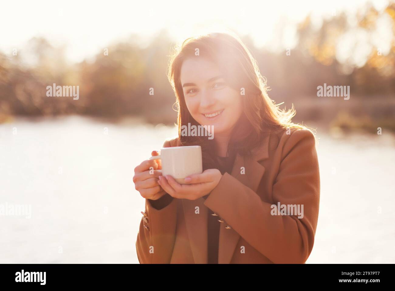 Smiling woman holding cup of tea near lake at sunset Stock Photo