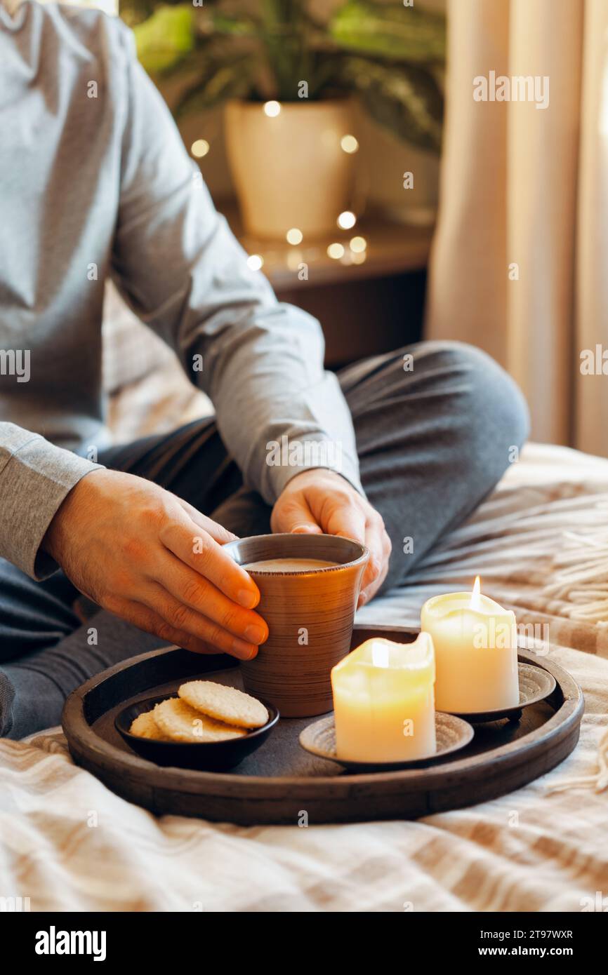 A caucasian man relaxing at home, lighting candle, drinking coffee in bed under blanket Stock Photo