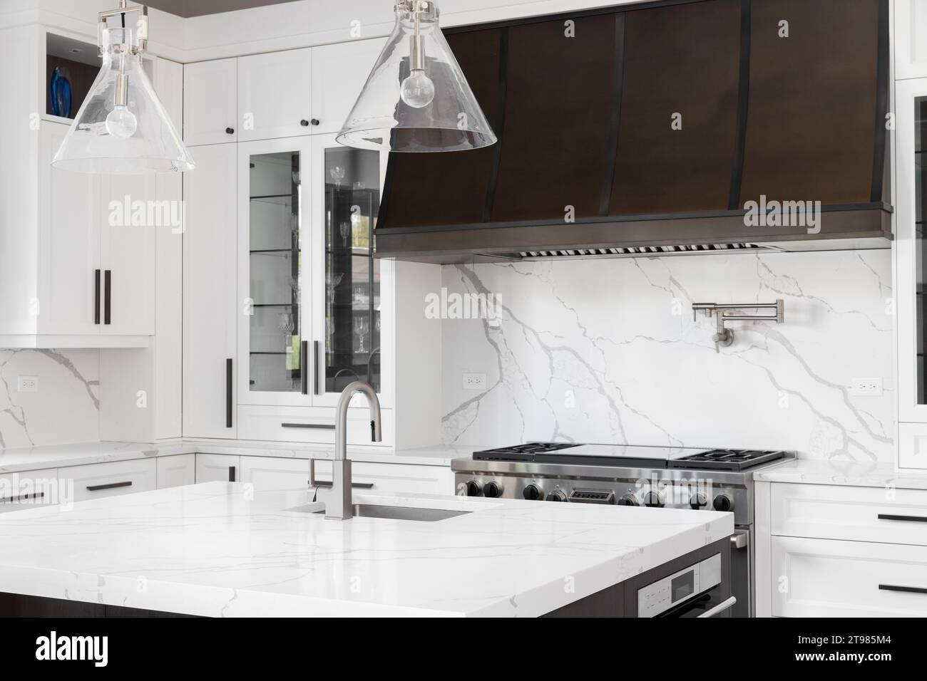 A kitchen detail with white cabinets, a brown metal range hood, white marble countertops and backsplash, and gold pendant lights. Stock Photo