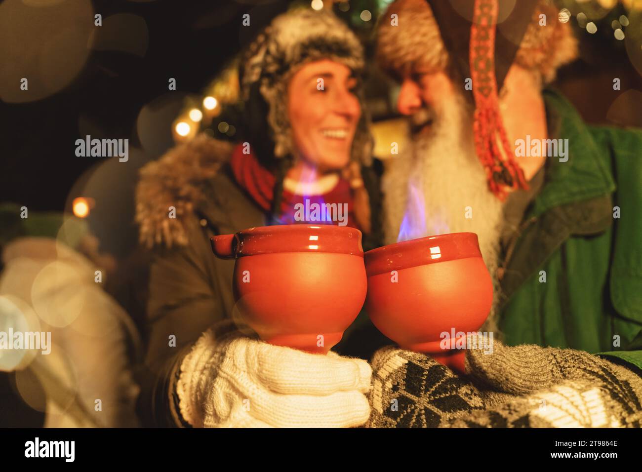 Joyful friends holding flaming fire tongs punch mugs at a Christmas market, glowing lights and bokeh in background. Stock Photo