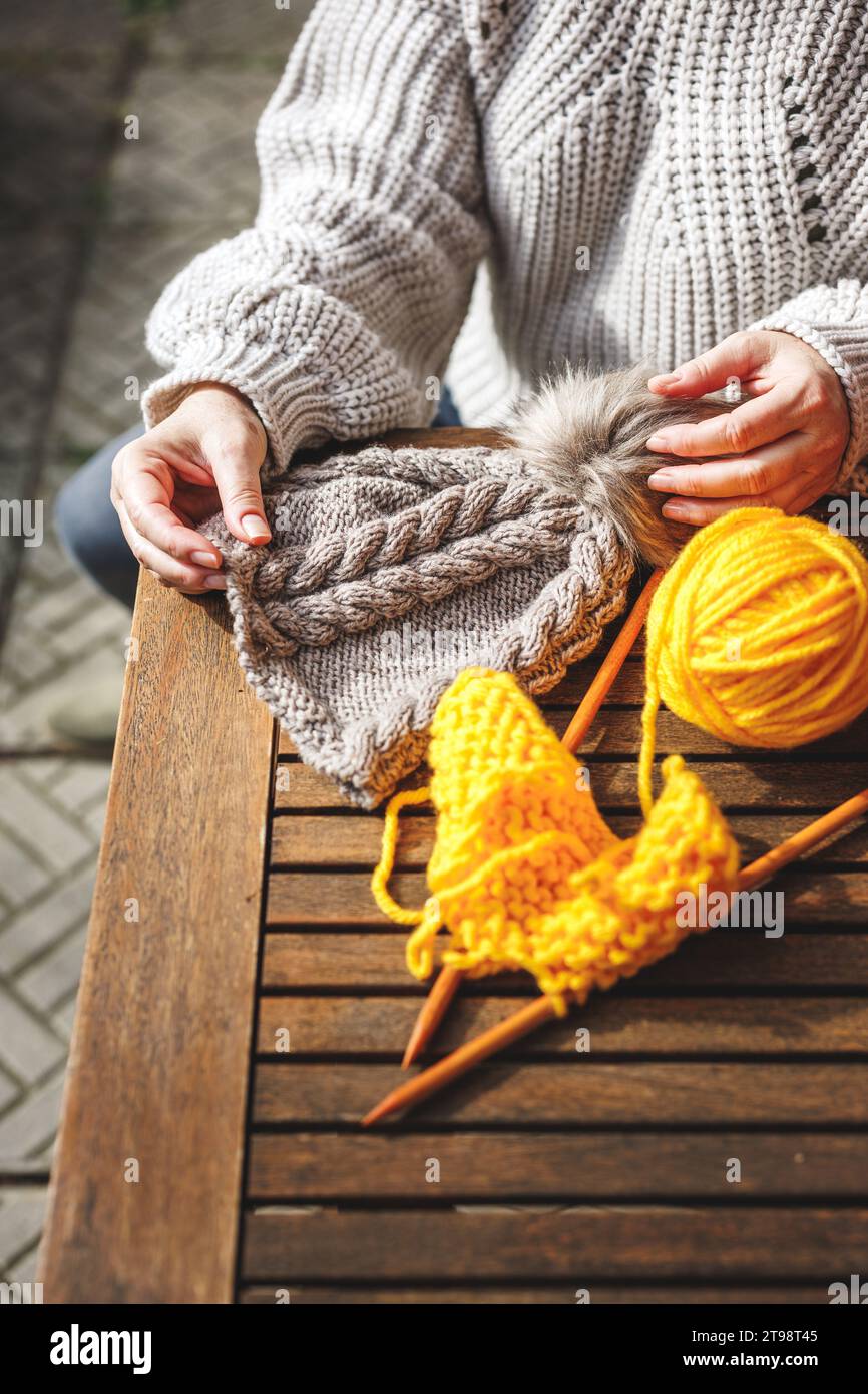 Woman holding handmade knit hat. Yellow ball of wool and knitting needle on table Stock Photo
