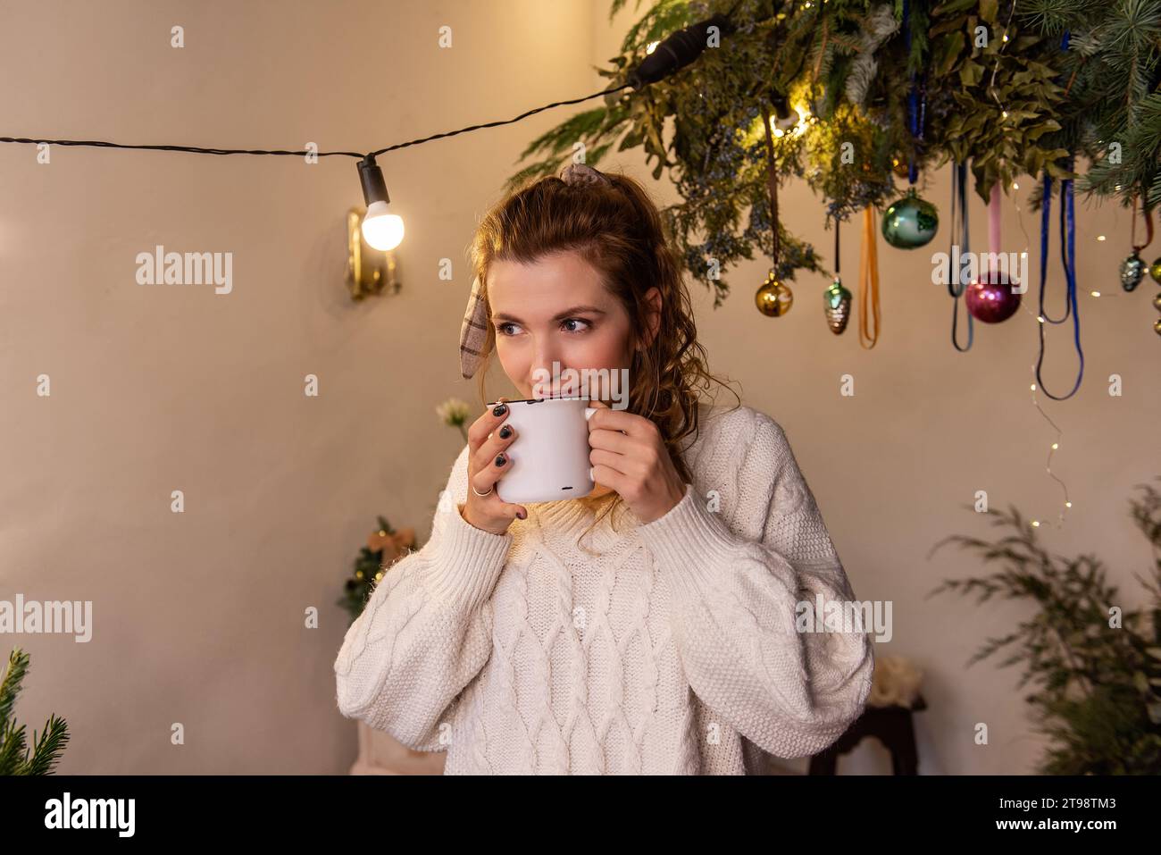 Close-up portrait of young woman holding white cup with hot drink. Girl drinks tea in Christmas interior in morning. Creative home decoration, hanging Stock Photo