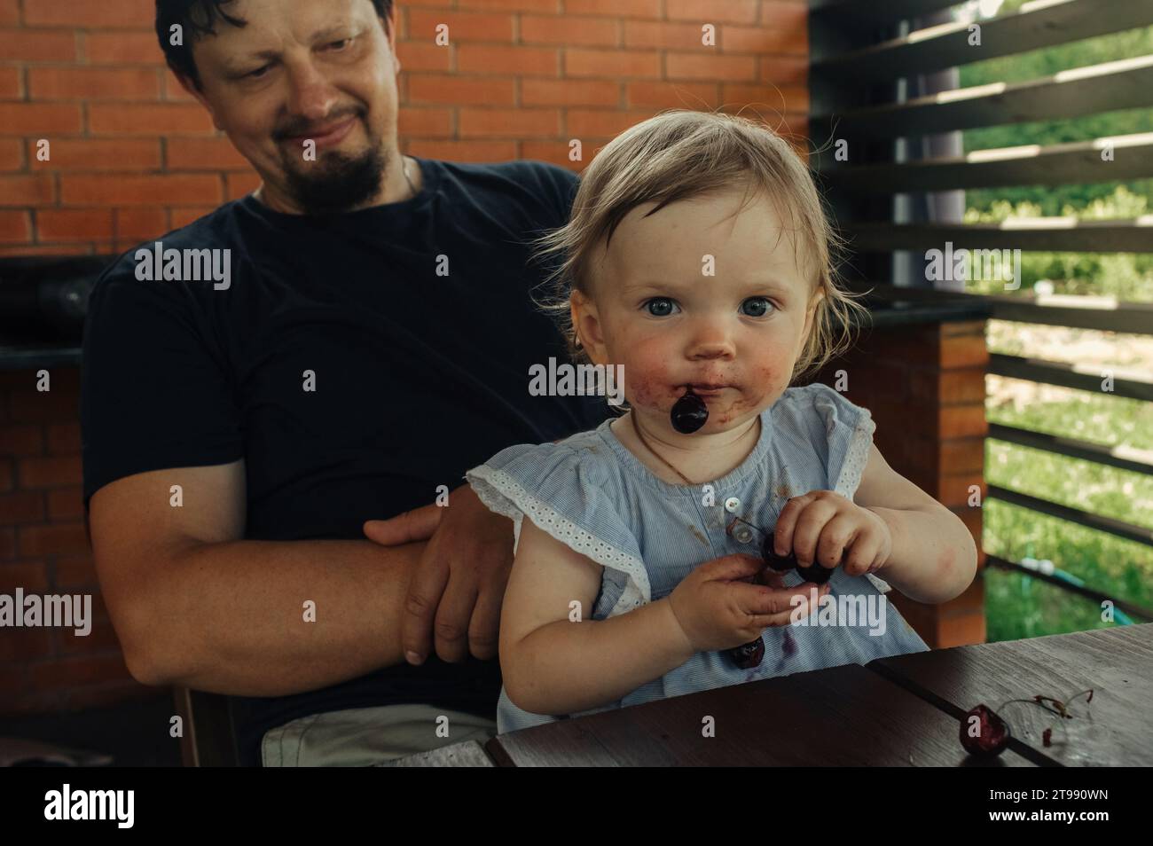 A little daughter sits in her father's arms and eats cherries. Stock Photo