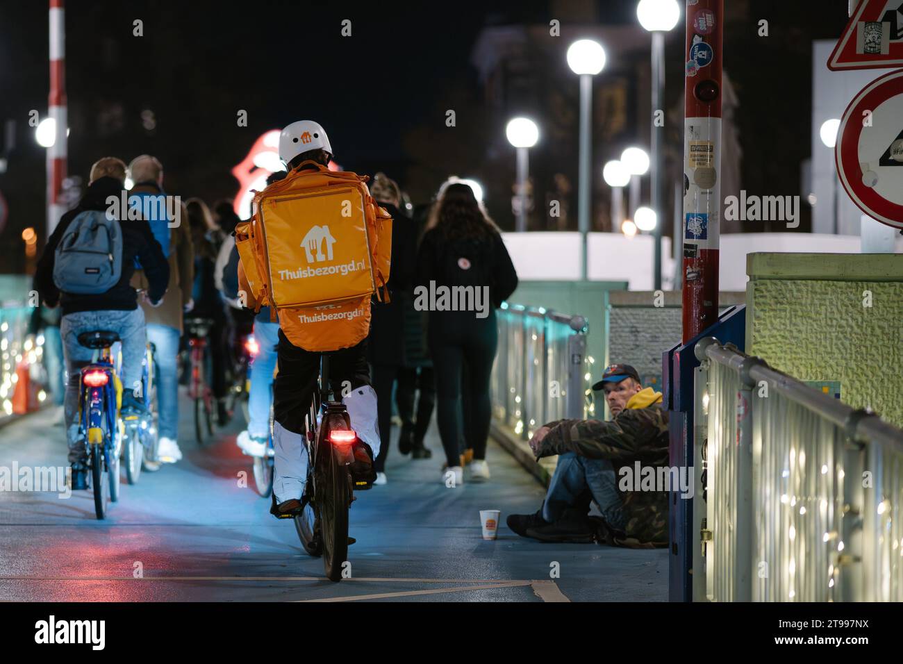 Groningen, Netherlands - 11 17 2023: Flash Delivery Person from Thuisbezorgd on Bicycle in a Big City Driving Past Begging Homeless Man Stock Photo