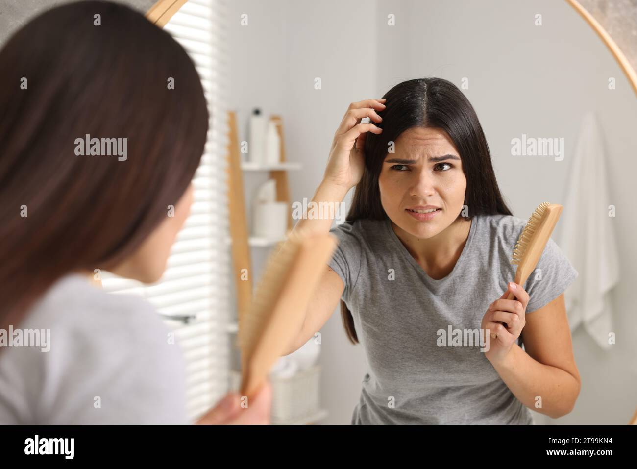 Emotional woman with brush examining her hair and scalp near mirror in bathroom. Dandruff problem Stock Photo