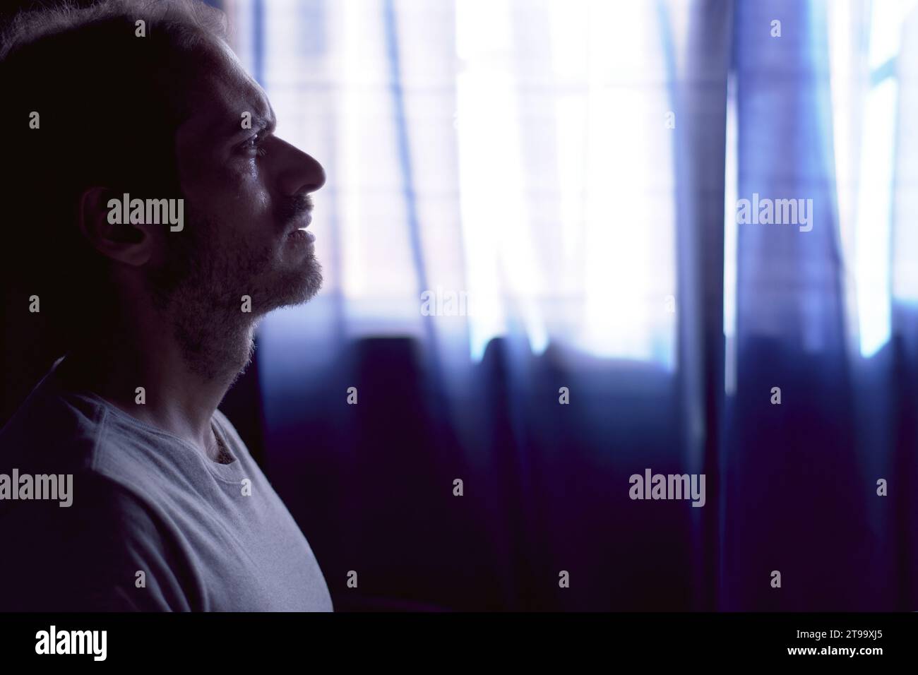 Depressed young bearded man crying in dark room. Dim light with window