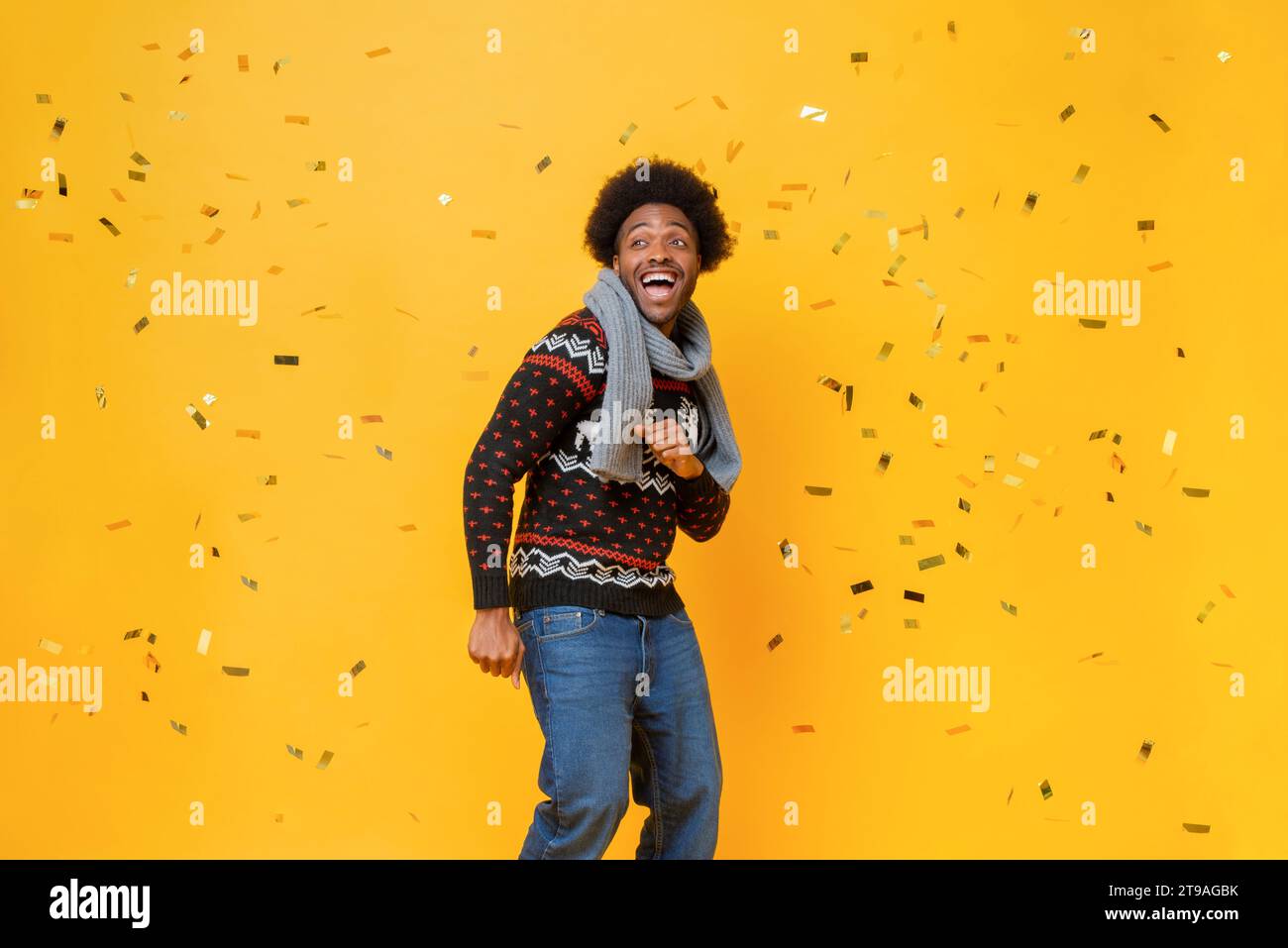 Festive studio shot portrait of cheerful smiling Afro African American man wearing Christmas sweater and scarf in yellow color background with confett Stock Photo