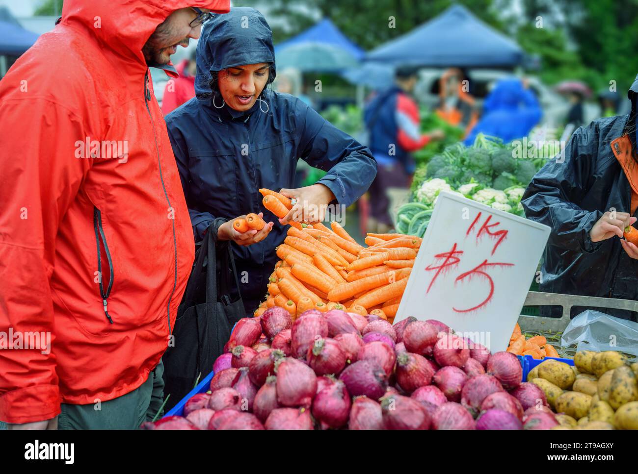 Couple choosing carrots in the market stalls in the rain. Unrecognizable people buying vegetables in the background. Stock Photo