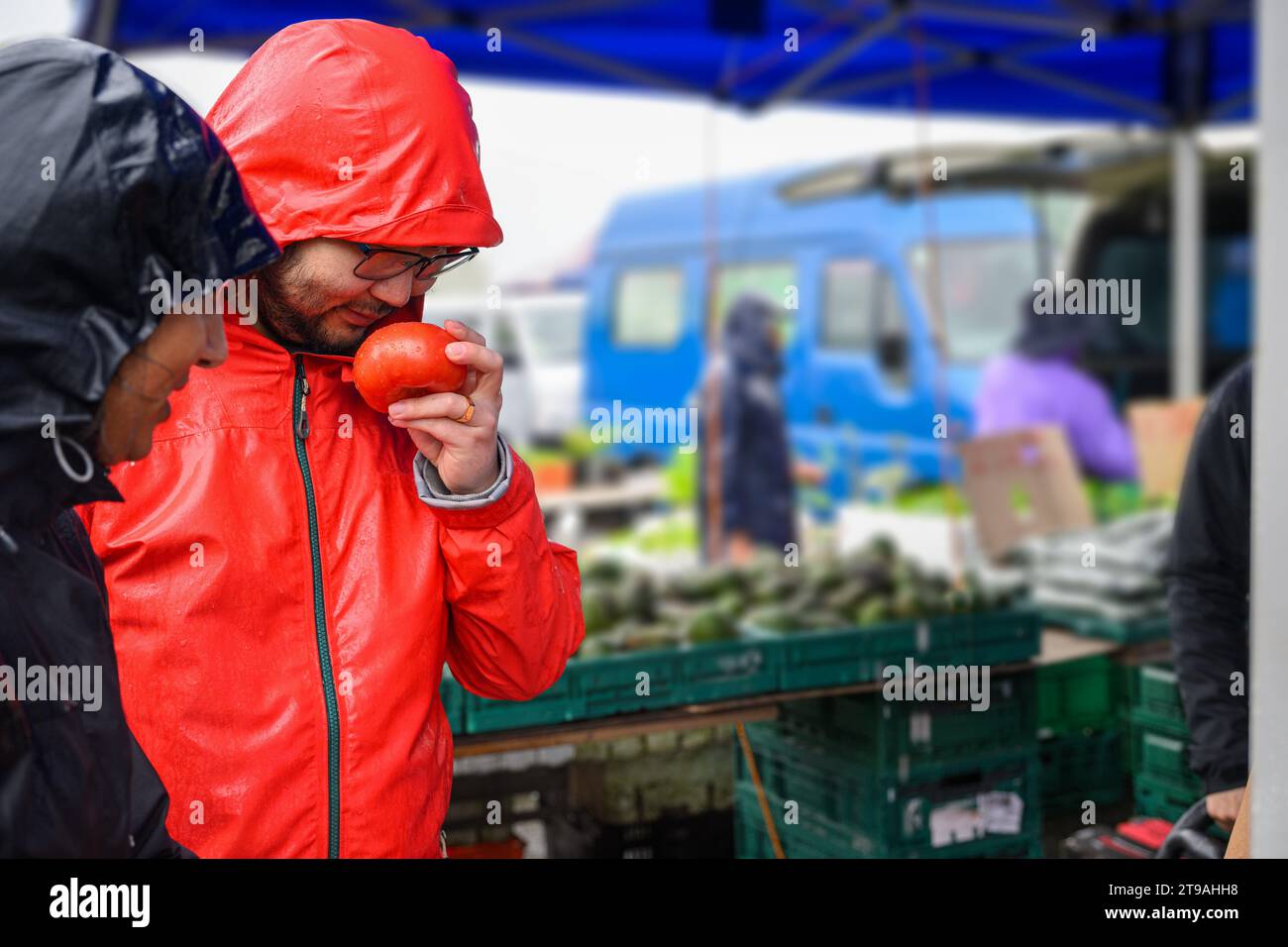 Man holding a ripe tomato and smelling its freshness at a vegetable market. Stock Photo