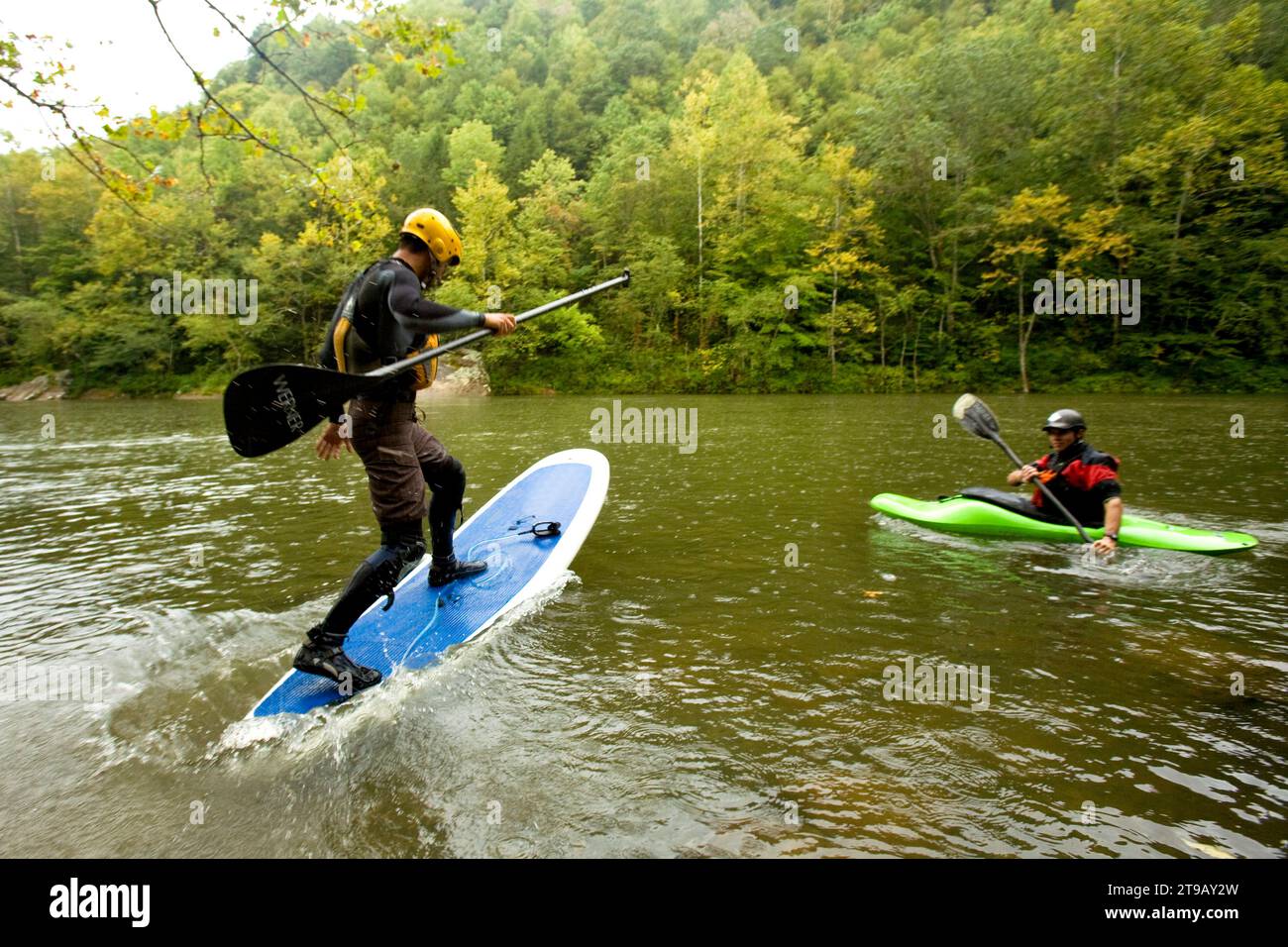 Male stand up paddleboarder jumping onto his board in a river while a kayaker watches. Stock Photo