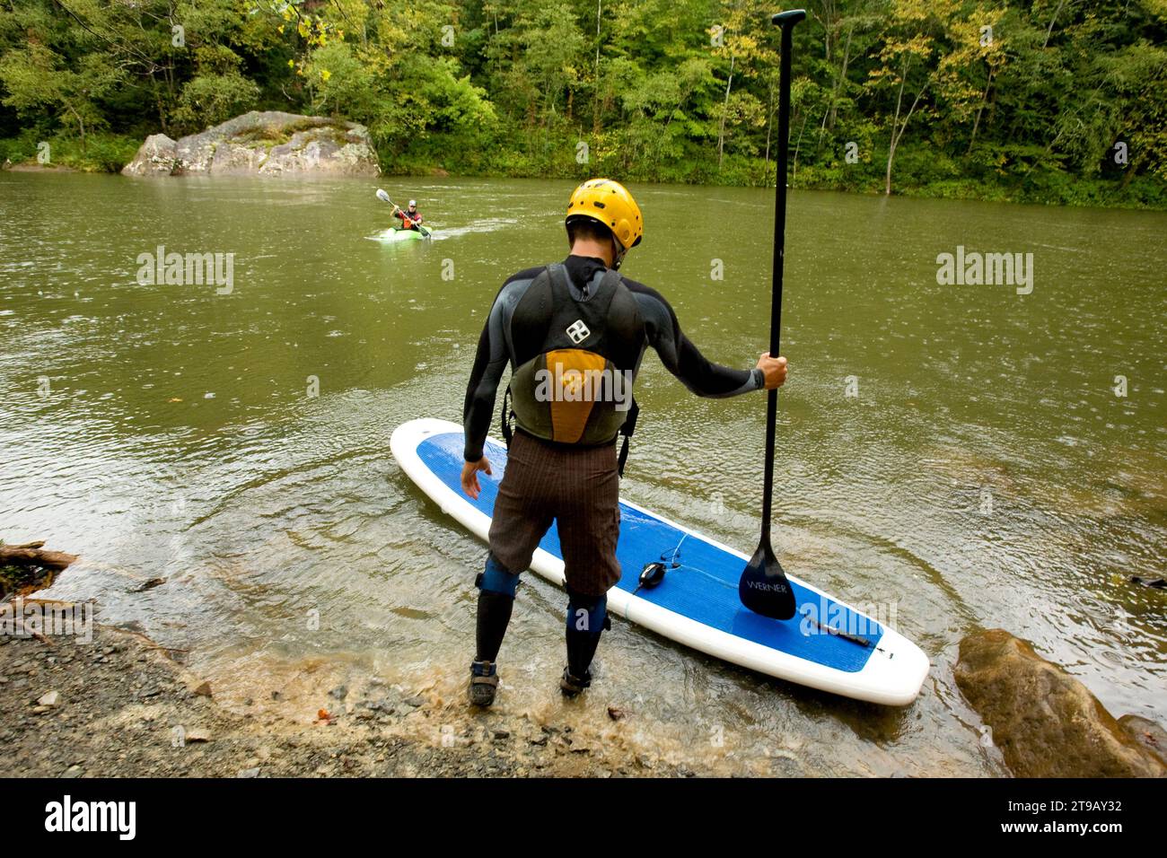 Male stand up paddleboarder getting ready to paddle a river while a kayaker watches from the river. Stock Photo