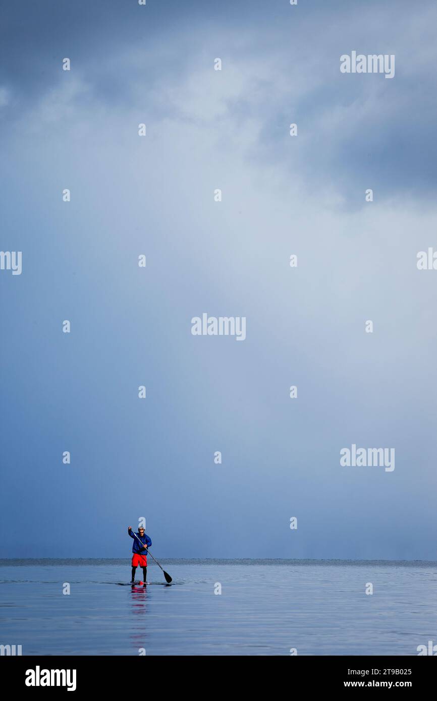 One man standup paddleboarding on a mountain lake in winter with a storm approaching. Stock Photo