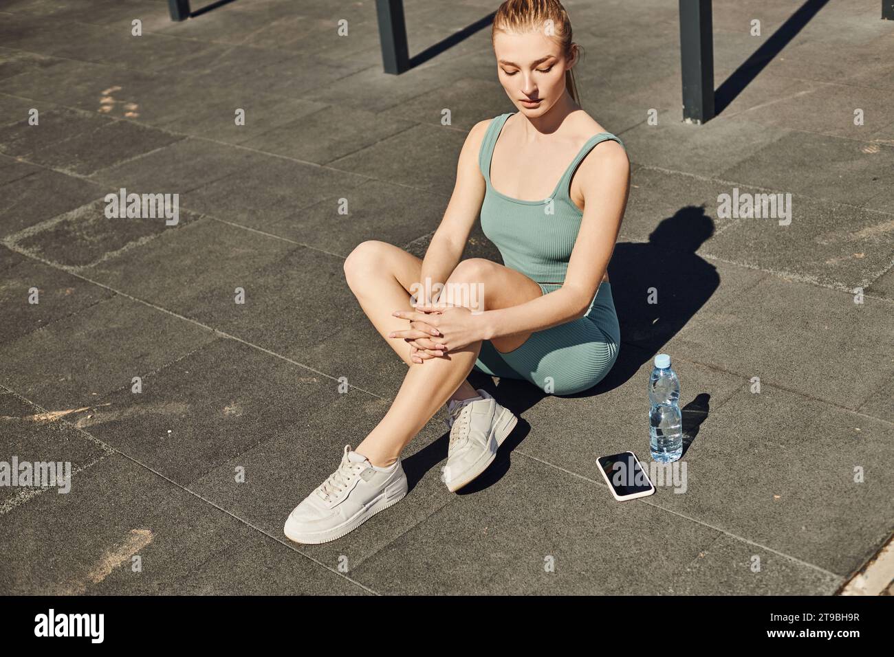 blonde sportswoman in tight activewear sitting next to bottle of water and smartphone on floor Stock Photo
