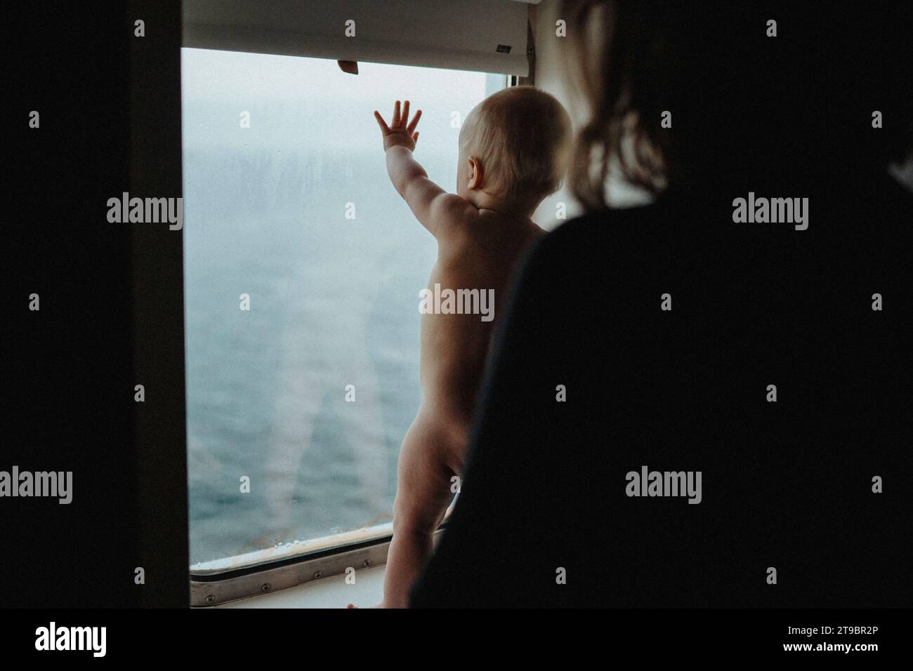 Mother supporting naked baby who is standing on window sill Stock Photo