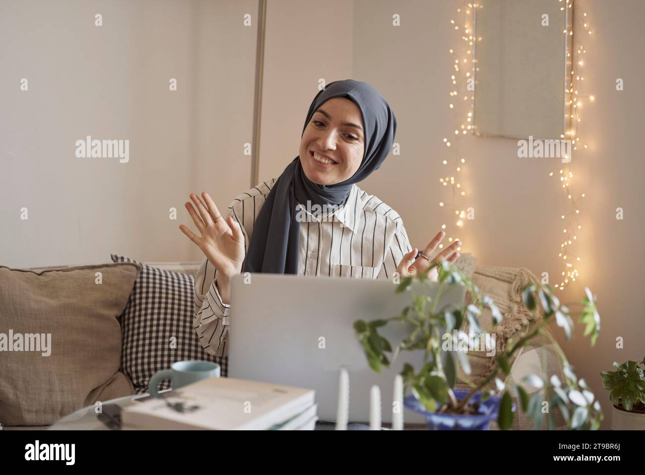 Smiling young businesswoman waving on video call over laptop while working in living room Stock Photo
