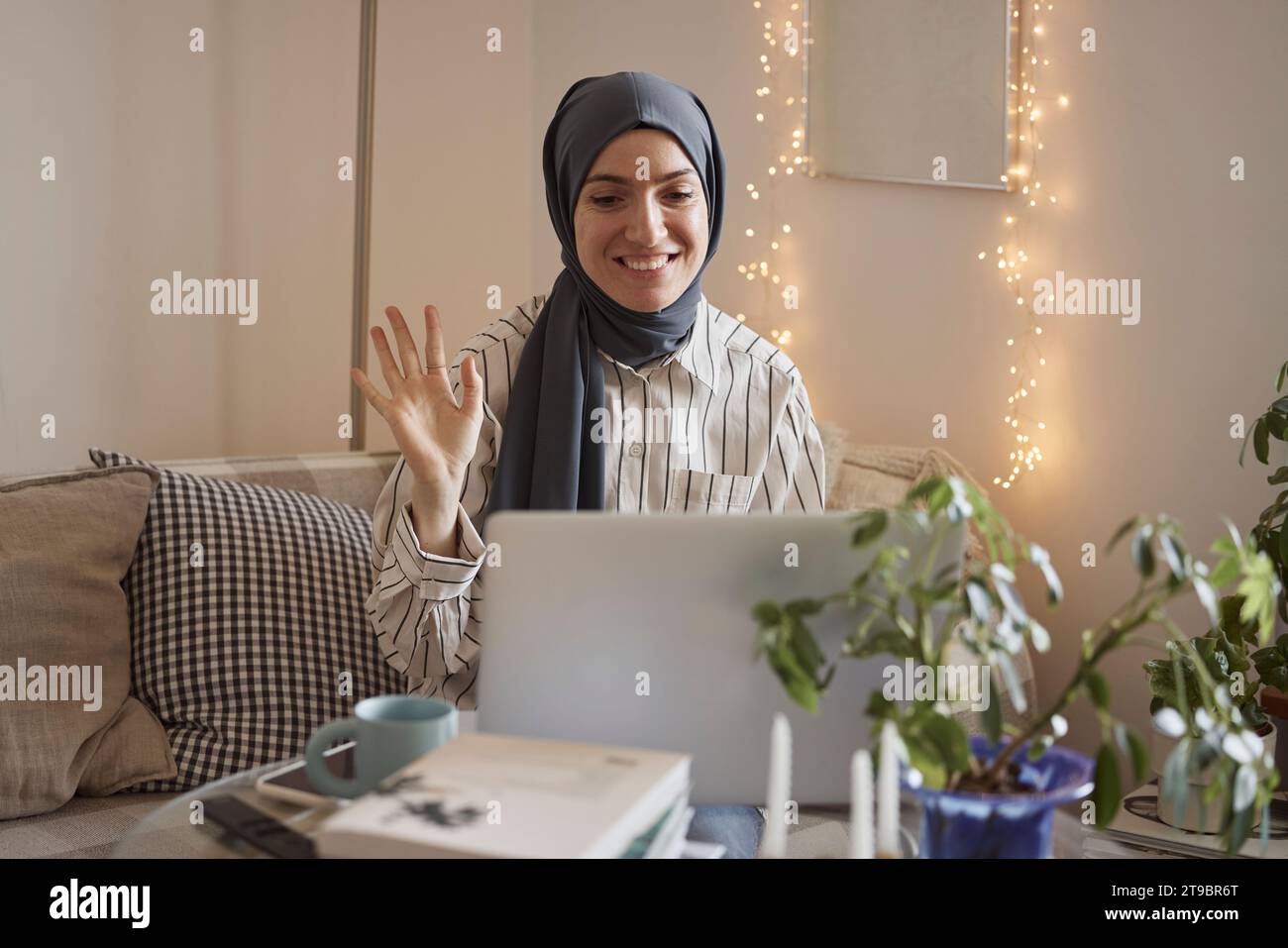 Happy female freelancer waving on video call over laptop while working in living room at home Stock Photo