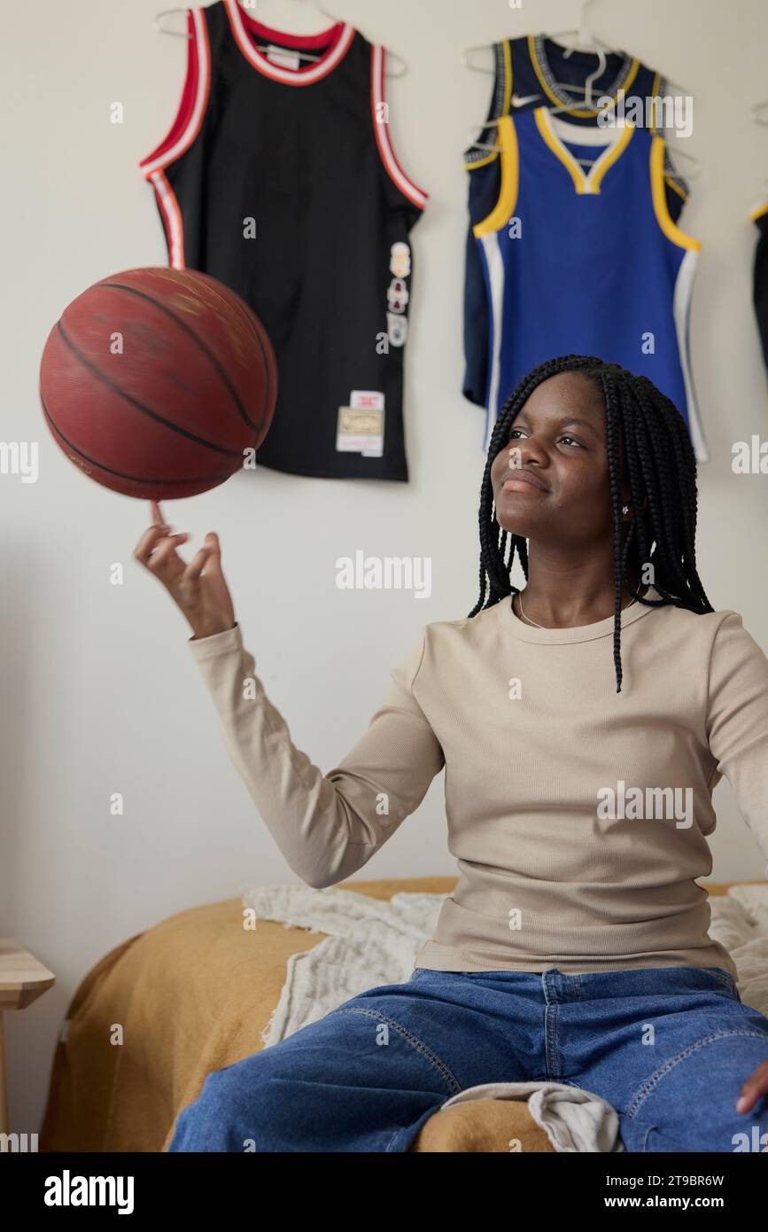 Teenage girl spinning basketball on her finger while sitting in bedroom at home Stock Photo