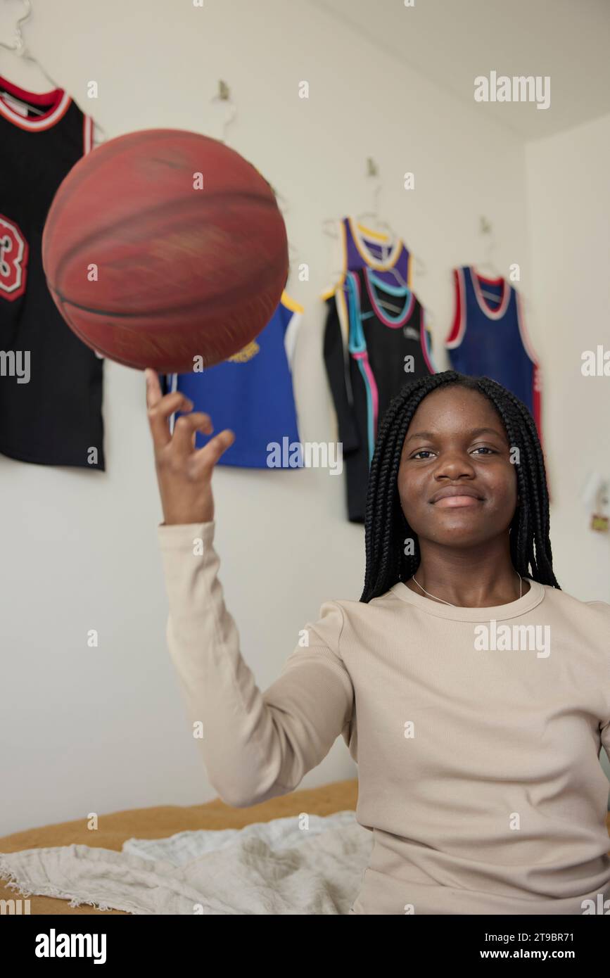 Portrait of teenage girl spinning basketball on her finger while sitting in bedroom Stock Photo