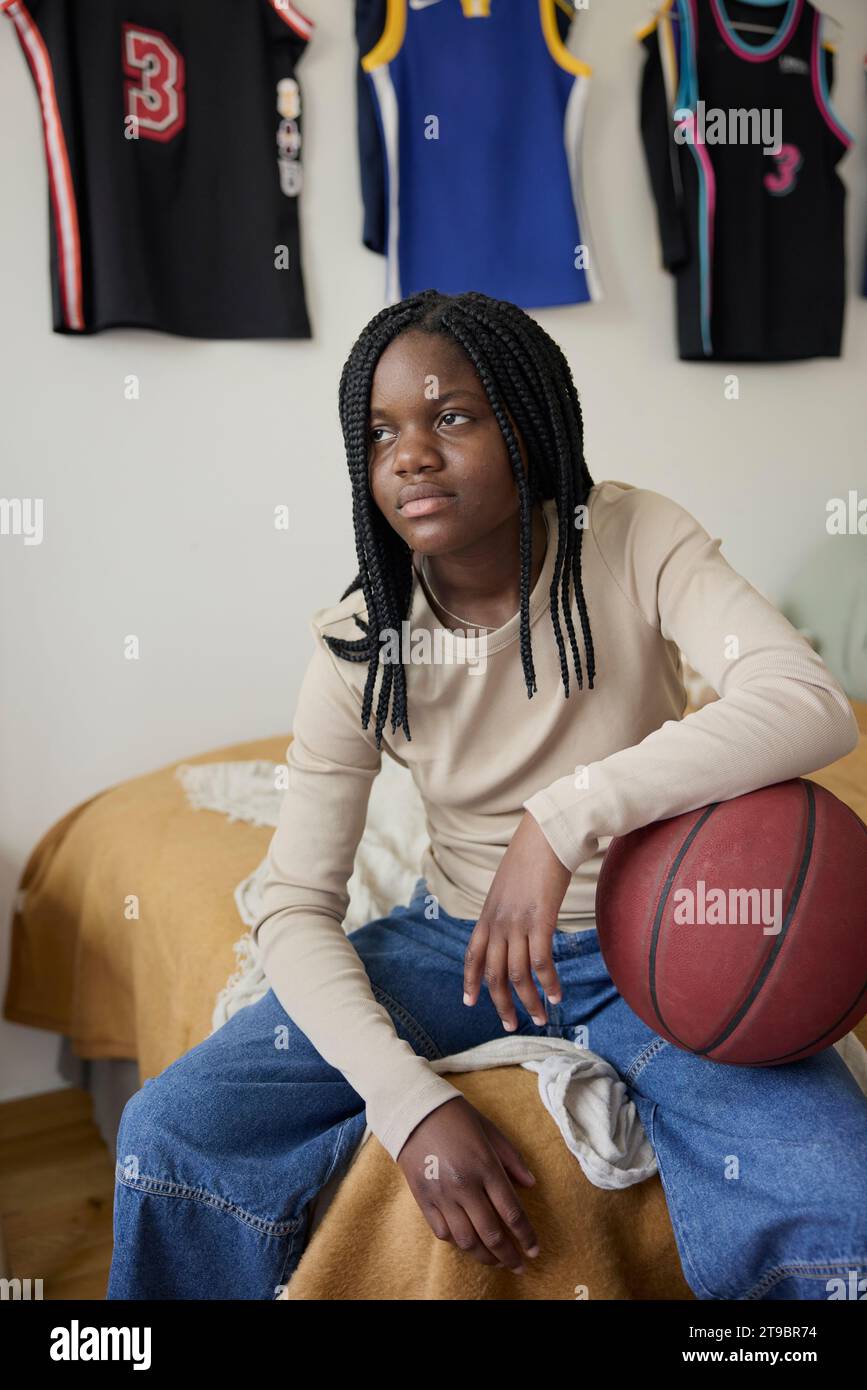 Contemplative female teenager with basketball sitting in bedroom Stock Photo