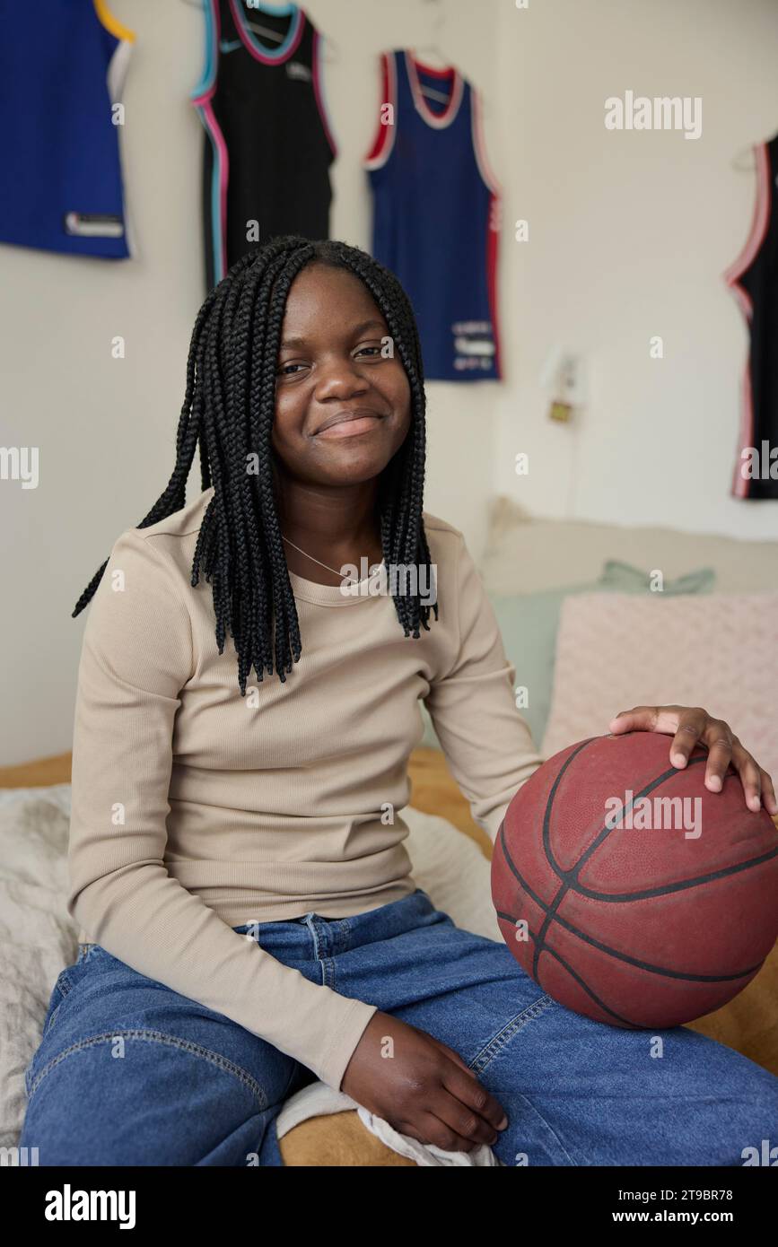 Smiling teenage girl with braided hair holding basketball while sitting in bedroom Stock Photo