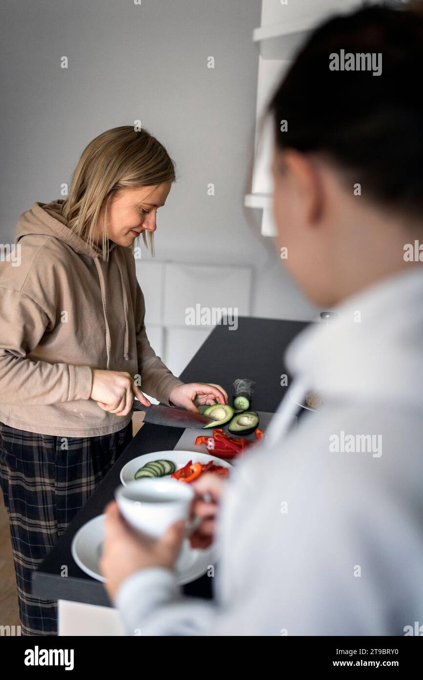 Smiling young woman cutting avocado at counter near girlfriend holding coffee cup in kitchen Stock Photo