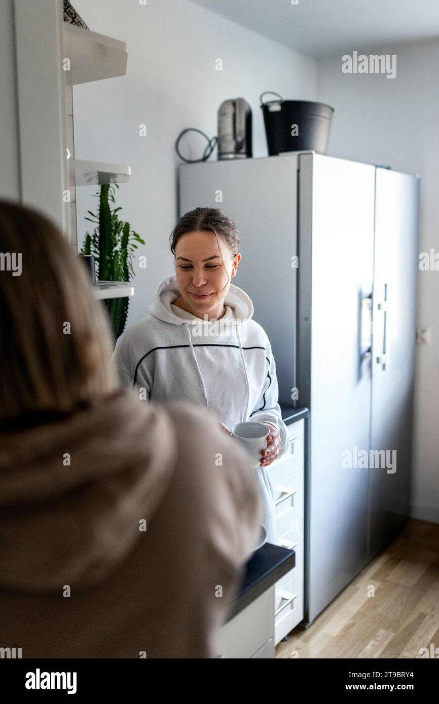 Smiling young woman holding coffee cup while standing near girlfriend in kitchen Stock Photo
