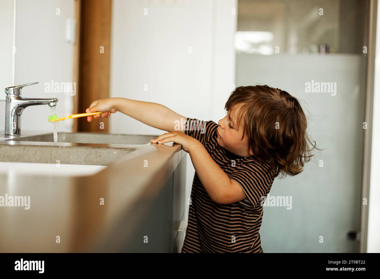 Boy holding toothbrush under running water from faucet Stock Photo