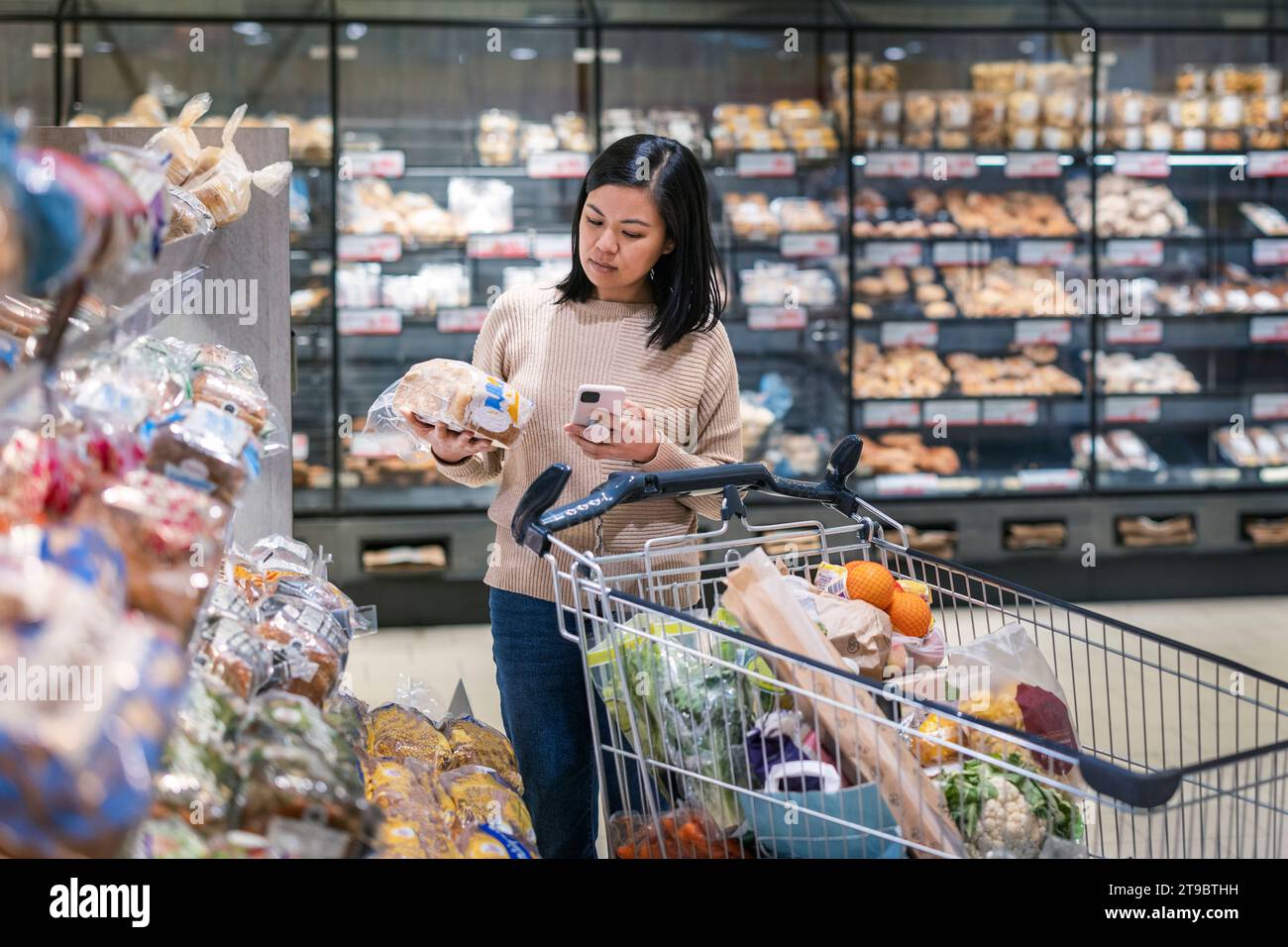Young woman using smart phone while holding package and shopping in supermarket Stock Photo