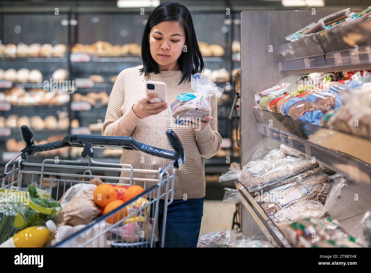 Young woman using smart phone while holding bread package and shopping in supermarket Stock Photo