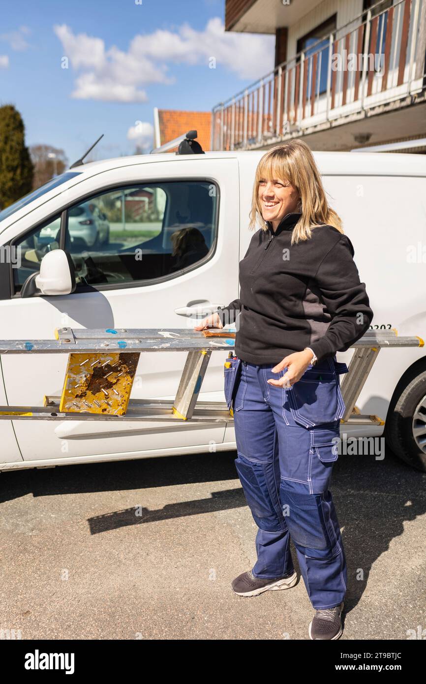 Smiling female technician carrying ladder while standing by van on road during sunny day Stock Photo