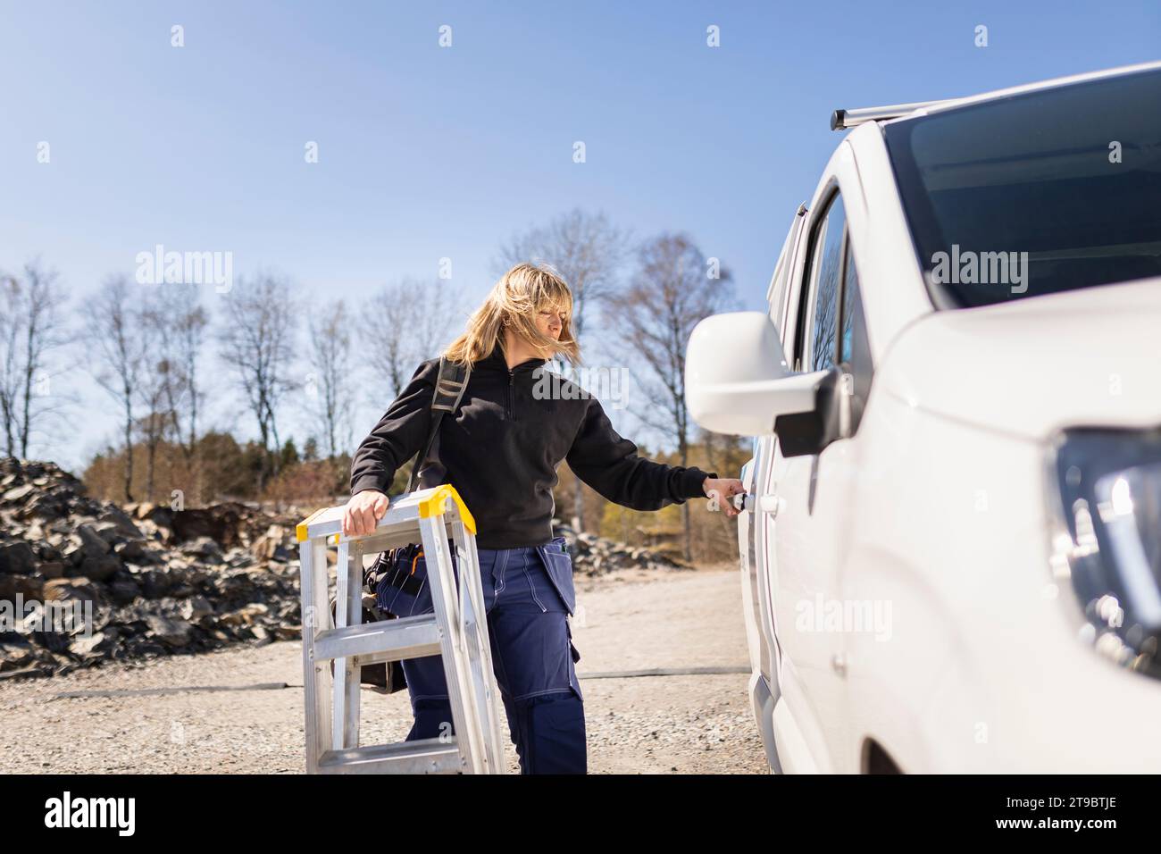 Female electrician with ladder opening van's door standing on sunny day Stock Photo