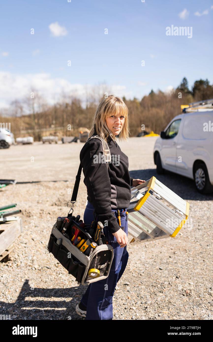 Female electrician carrying ladder and toolbox standing on road Stock Photo