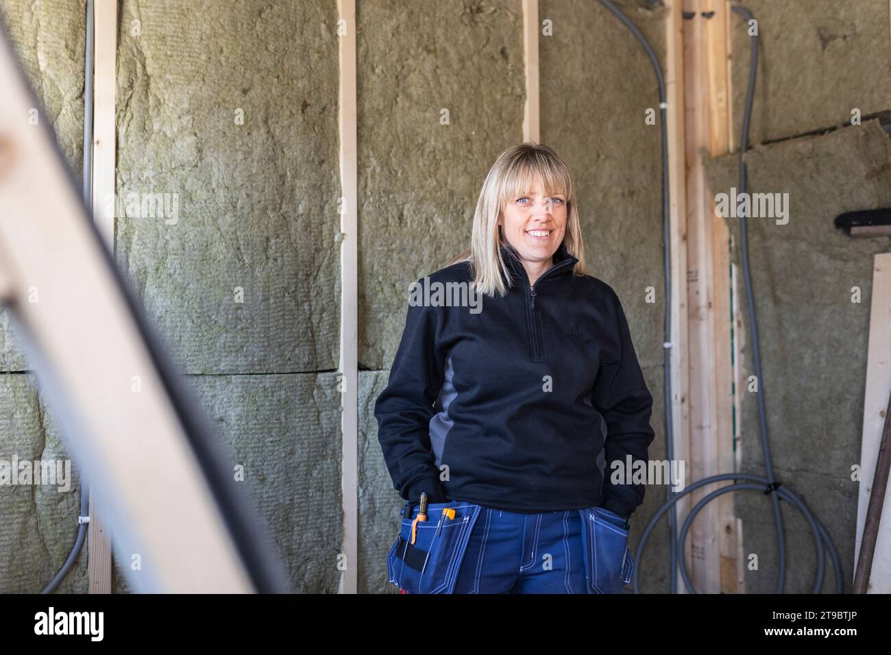 Smiling female blue-collar worker with hands in pockets standing against wall Stock Photo
