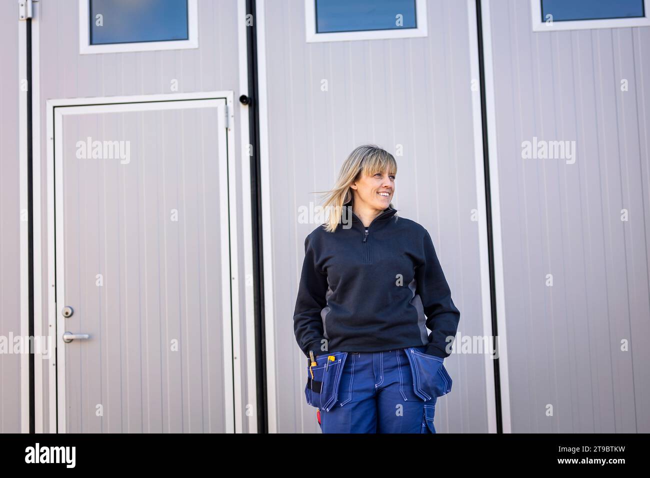 Female blue-collar worker with hands in pockets standing against door Stock Photo