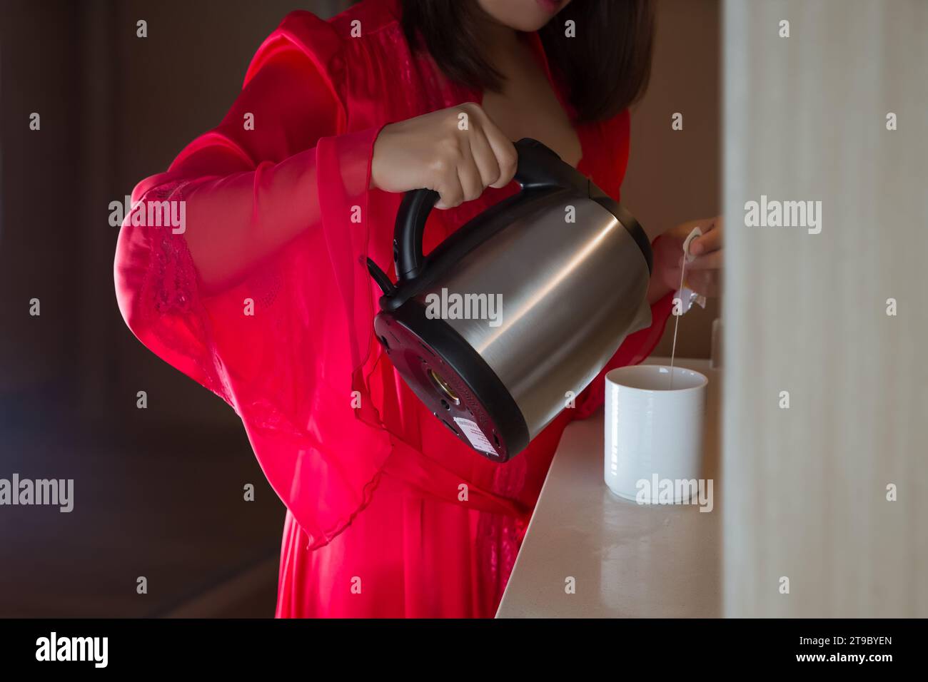 A woman in pajamas and a red satin robe stand pouring hot water to prepare tea at night. Stock Photo