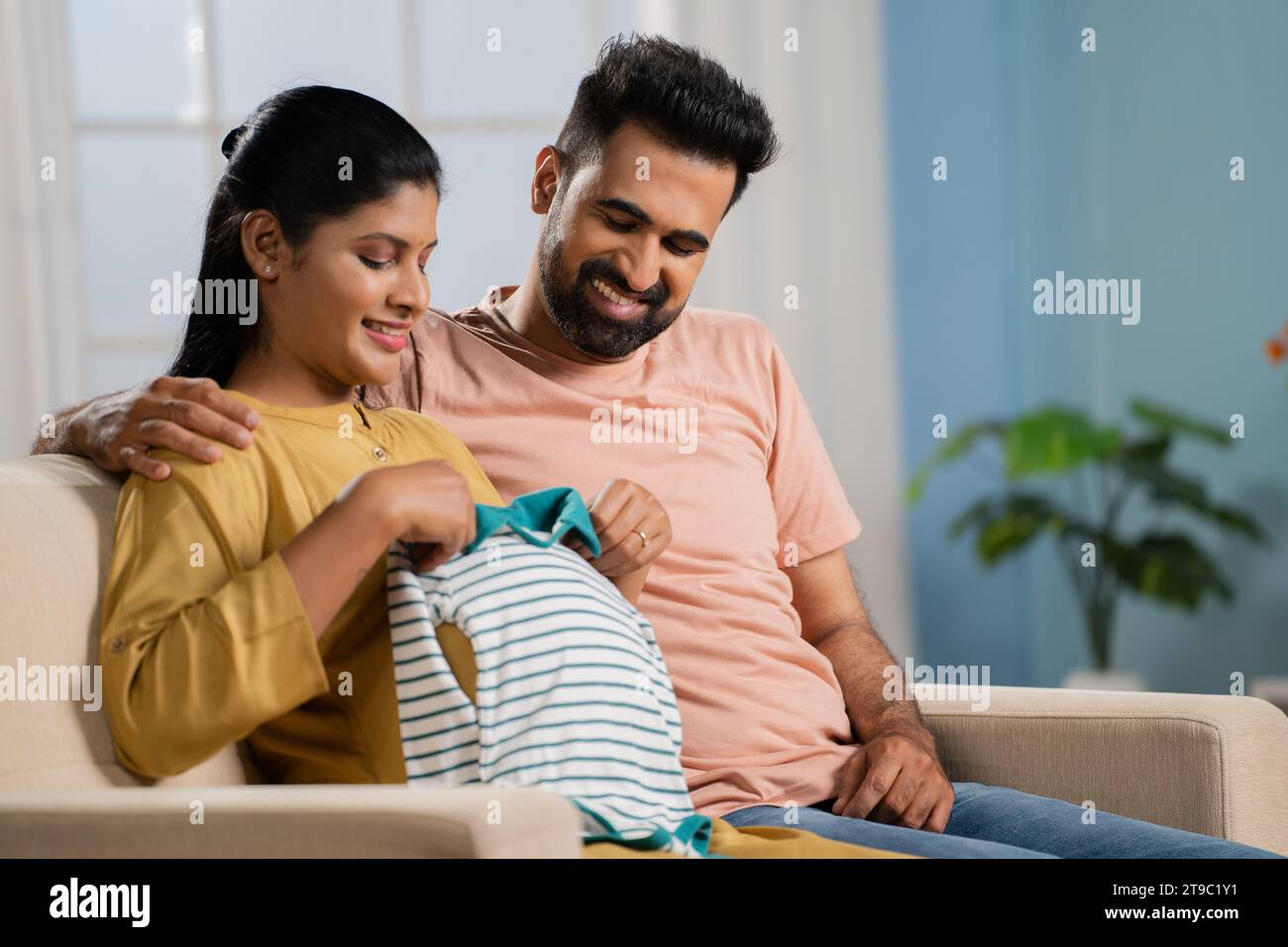 Happy Indian Pregnant woman checking baby dress by placing on tummy after given by husband - concept Parenthood journey, Expectant couples and bonding Stock Photo