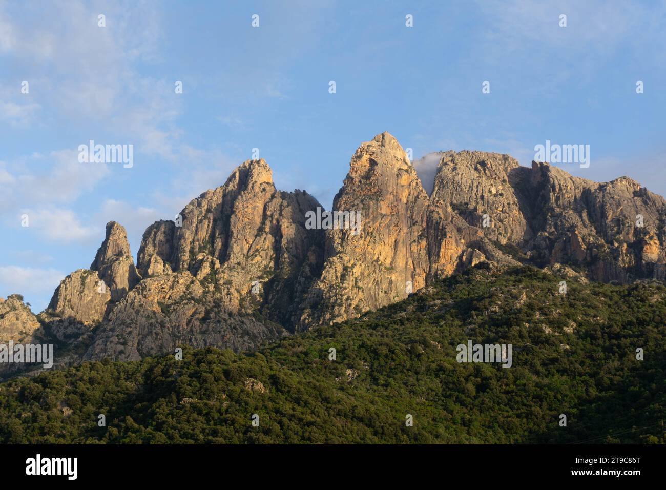 Golden hour on Corsica unveils rocky orange peaks adorned with lush greenery against a backdrop of a serene blue sky. Corsica, mountains, France. Stock Photo