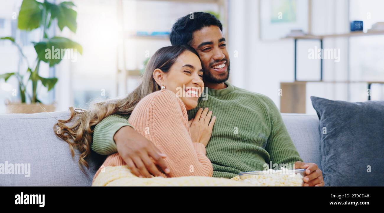 Love, hug and couple on watching tv on a sofa with popcorn for movie, film or streaming show at home. Relax, television and people embrace in a living Stock Photo