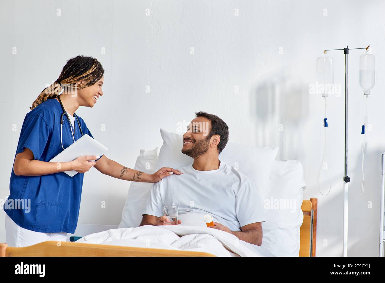 jolly african american nurse smiling happily at her indian patient that lying in hospital bed Stock Photo