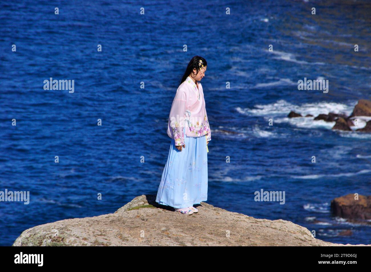 The delicate colours and materials of the traditionally dressed oriental lady contrasts with the rugged granite rocks and bright blue sea near Land Stock Photo