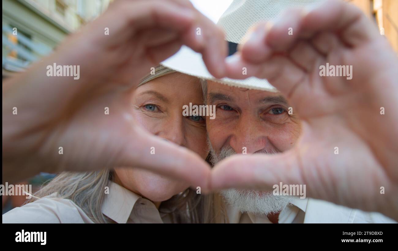 Close up portrait happy Caucasian woman man old retired family couple looking at camera through joined fingers heart gesture love feelings celebrate Stock Photo