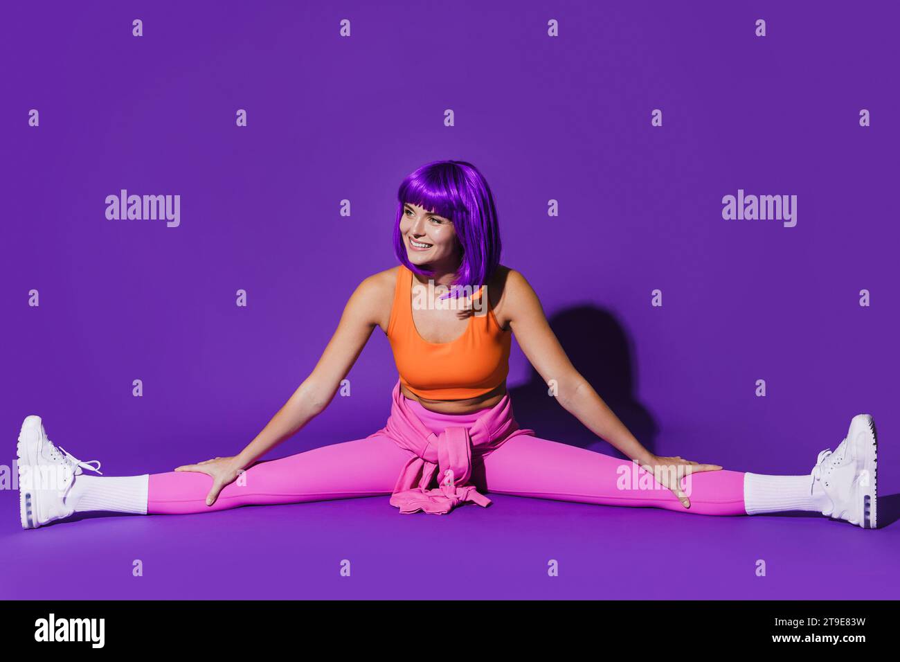 Young cheerful woman wearing colorful sportswear doing splits against purple background Stock Photo