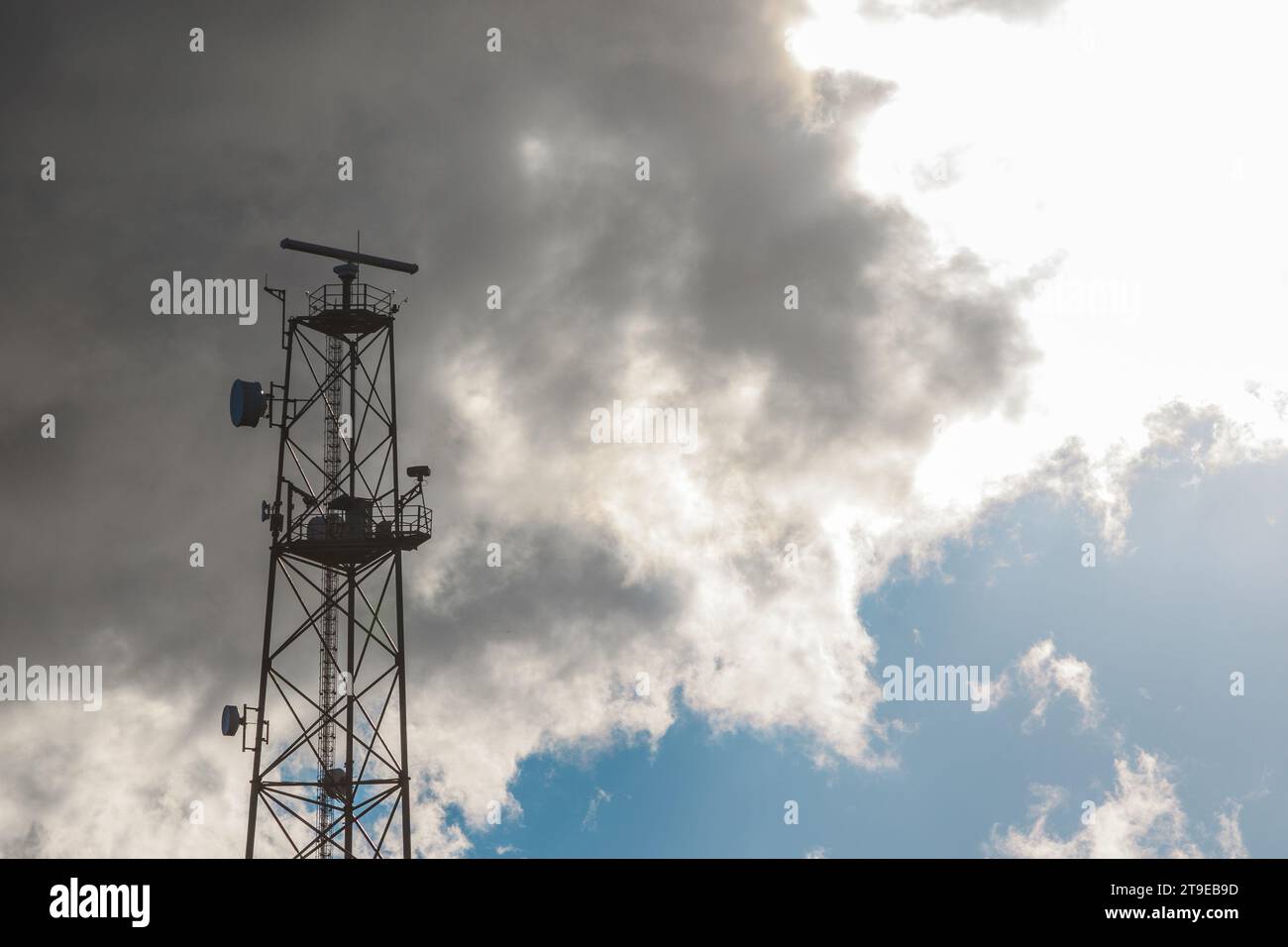 Upward shot of top of a metallic telecommunication tower on a sky background with large white cloud. Stock Photo