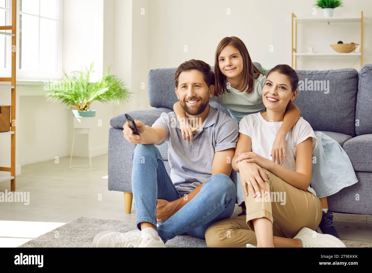 Happy mom, dad and child sitting on floor by sofa and watching television show together Stock Photo