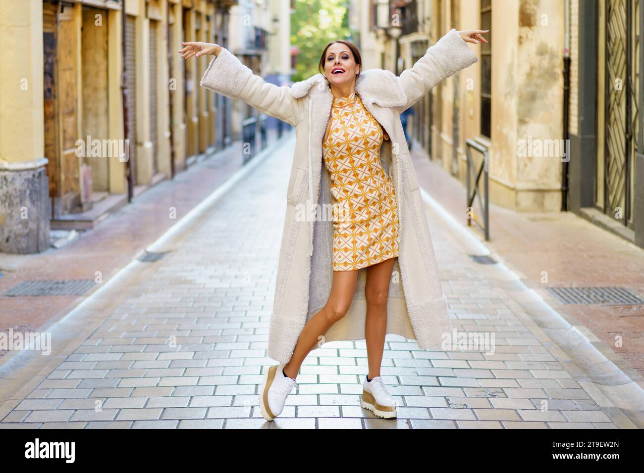 Stylish young woman wearing fashionable long coat with dress and shoe dancing on street against building background Stock Photo
