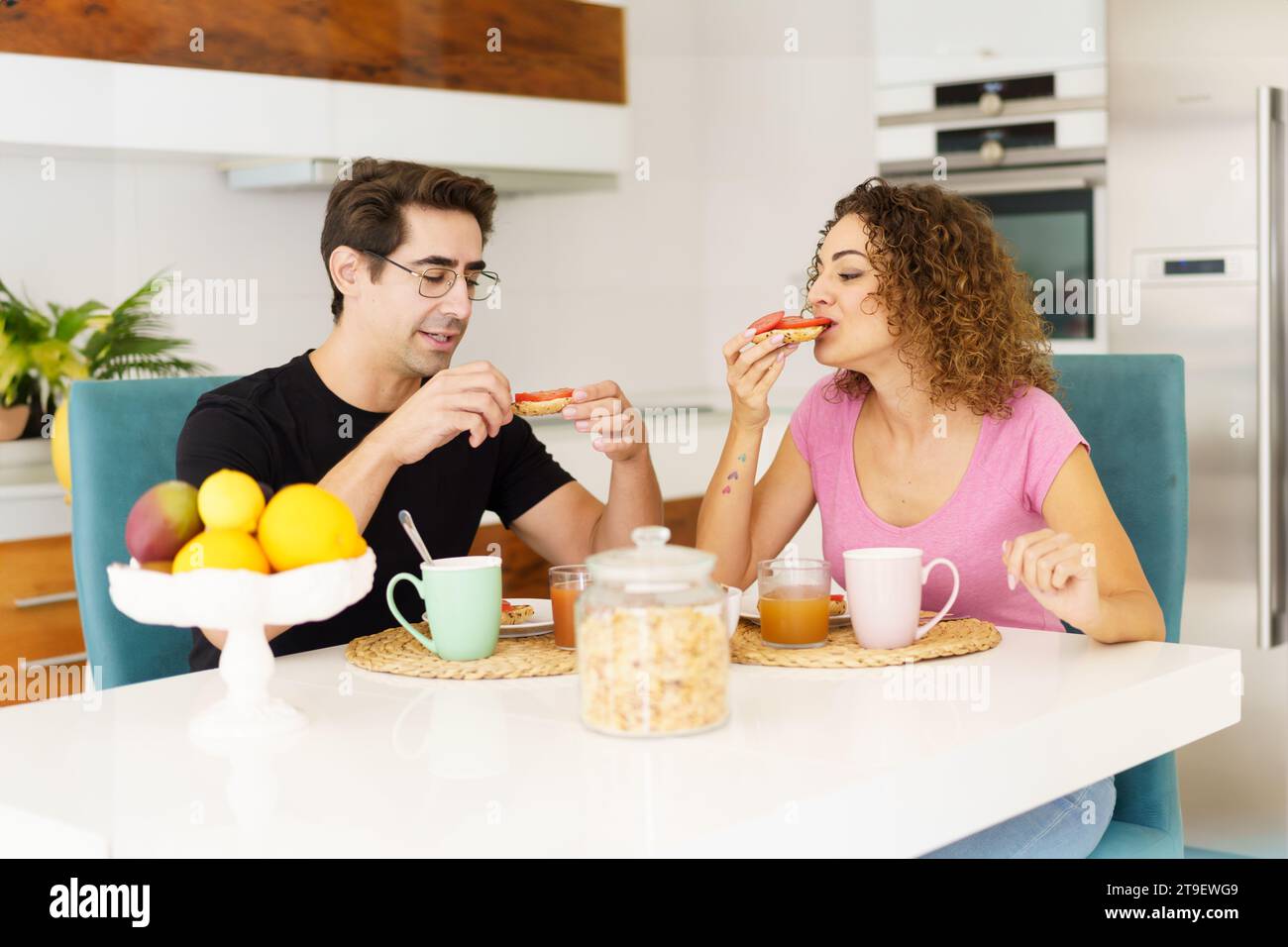 Happy adult couple in conversation and looking down while sitting at dining table in kitchen with juice in glasses, on placemat near fruits in bowl an Stock Photo