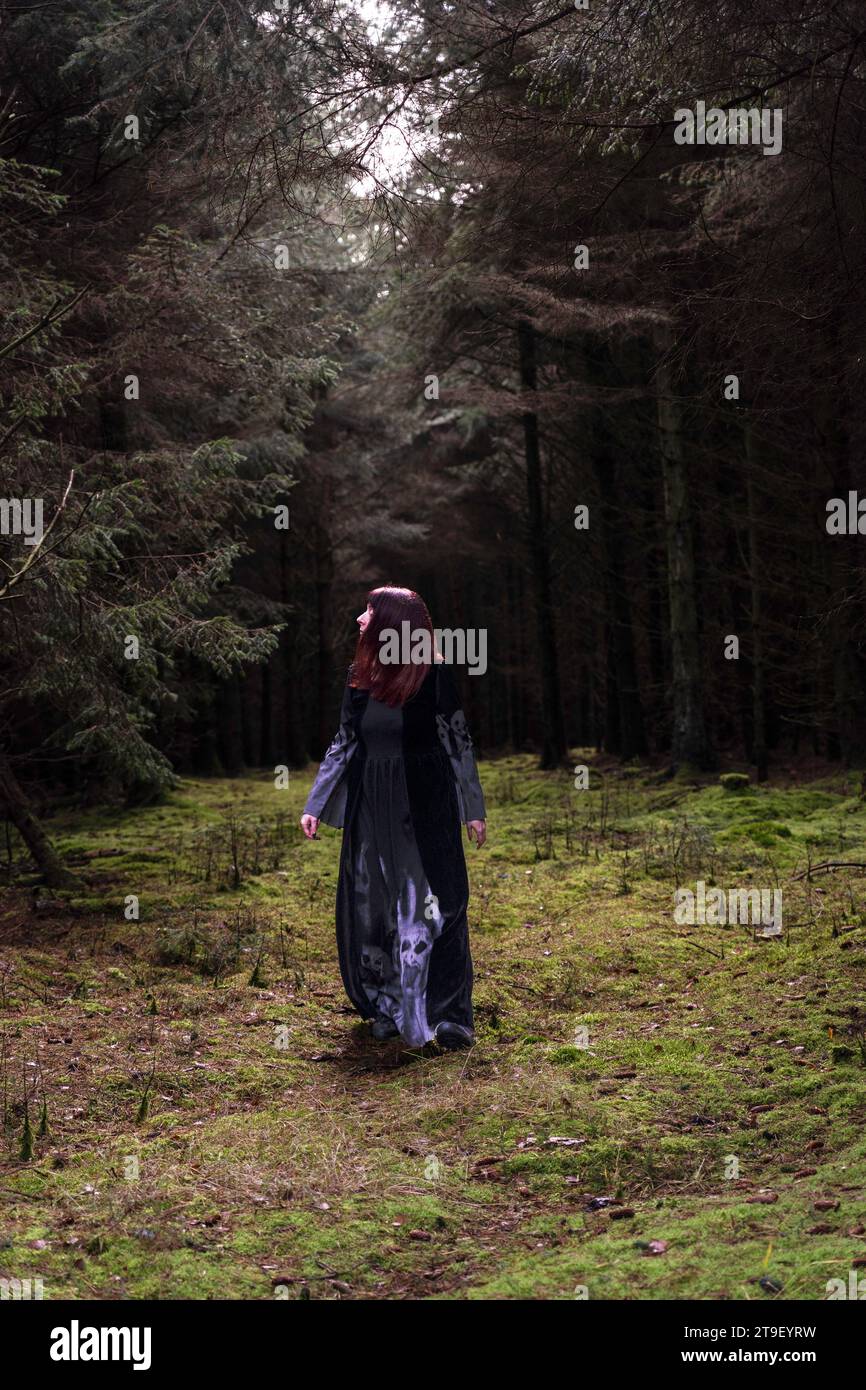 woman in black and grey dress with bat sleeves and skulls walking towards camera in a forest Stock Photo