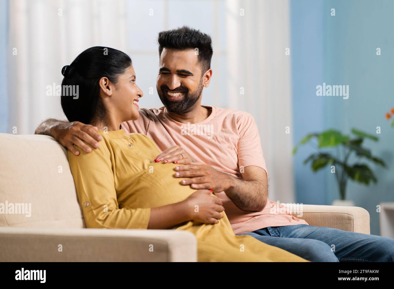 Happy Indian husband showing laptop to pregnant wife at home - concept of parenthood, cyberspace and surfing internet. Stock Photo