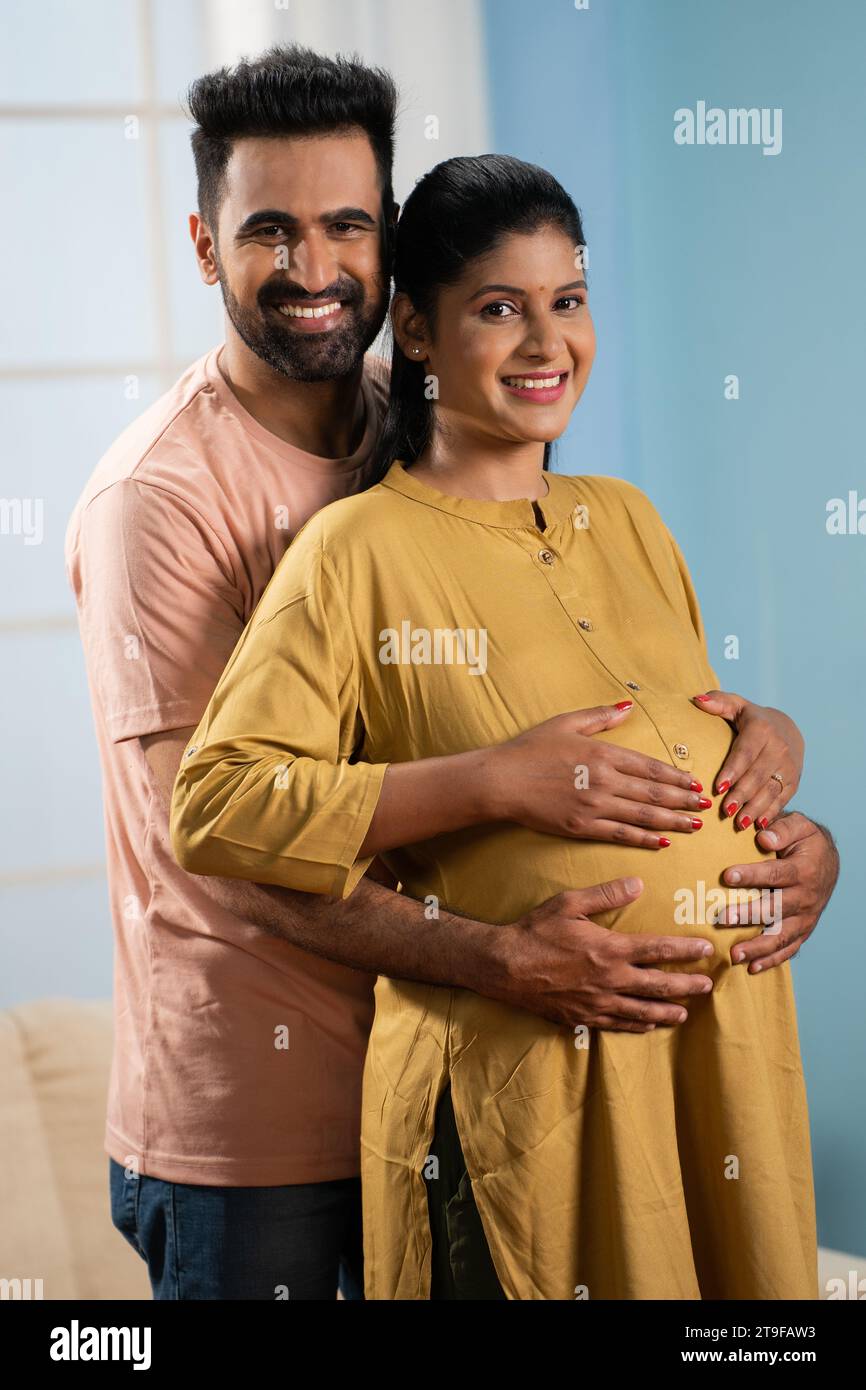 vertical Portrait shot of happy indian Pregnant woman with husband looking at camera while talking at home - concept of parenthood, family support and Stock Photo