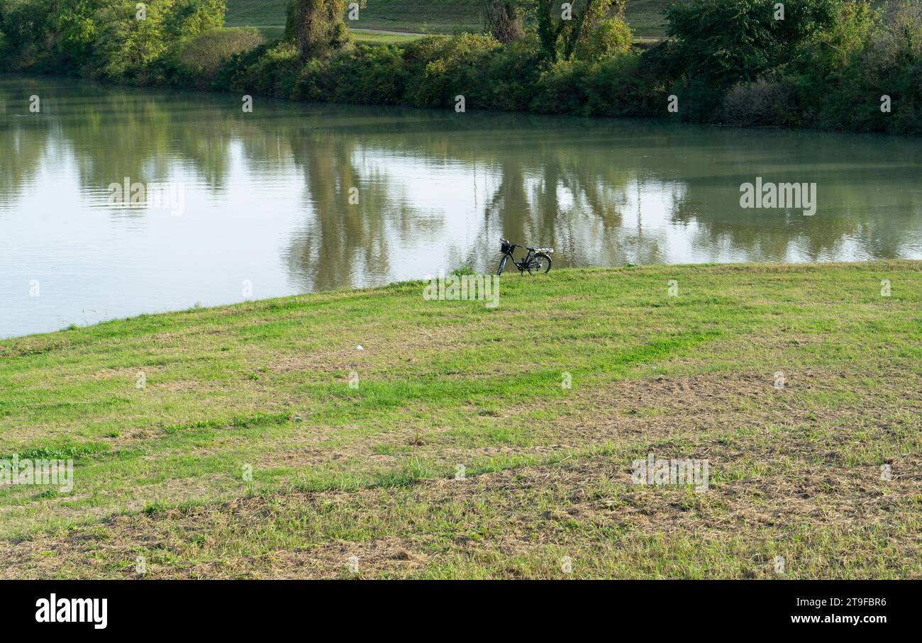 A bicycle parked on the bank of a river during an ecological nature-oriented intinerary. Green grass and trees in the background contribute to the ser Stock Photo